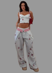 A woman modeling a white two-piece outfit featuring a cropped camisole top and matching wide-leg pants, both decorated with pink floral prints. She is wearing a pink waist tie belt and carrying a red shoulder bag, standing on a wooden parquet floor against a white wall.