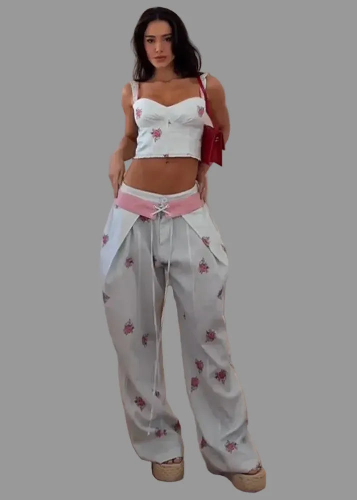 A woman modeling a white two-piece outfit featuring a cropped camisole top and matching wide-leg pants, both decorated with pink floral prints. She is wearing a pink waist tie belt and carrying a red shoulder bag, standing on a wooden parquet floor against a white wall.