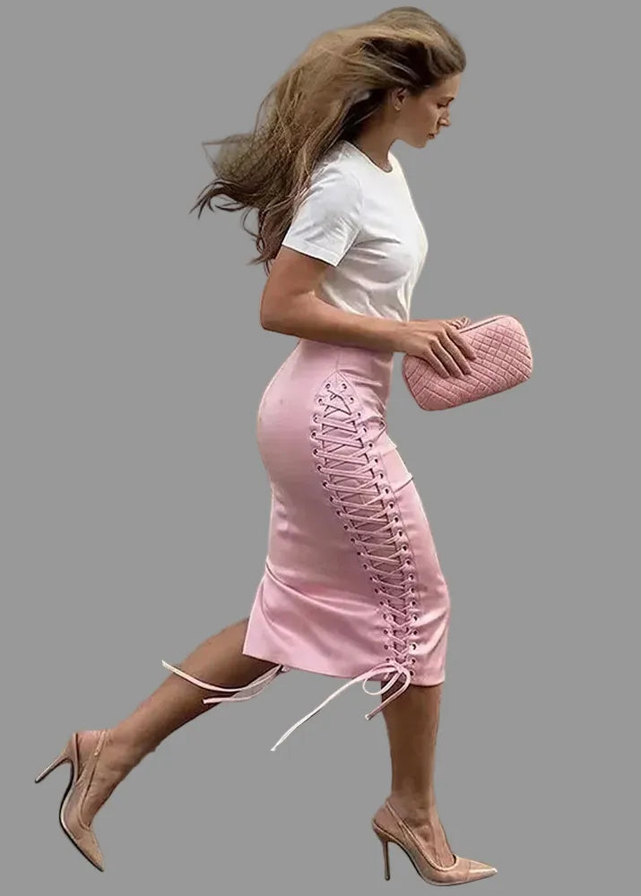 A woman walking outdoors wearing a fitted light pink midi skirt with side lace-up detailing, paired with a white short-sleeve top and nude heels. She is holding a pink quilted clutch, with a stone wall and pavement in the background.
