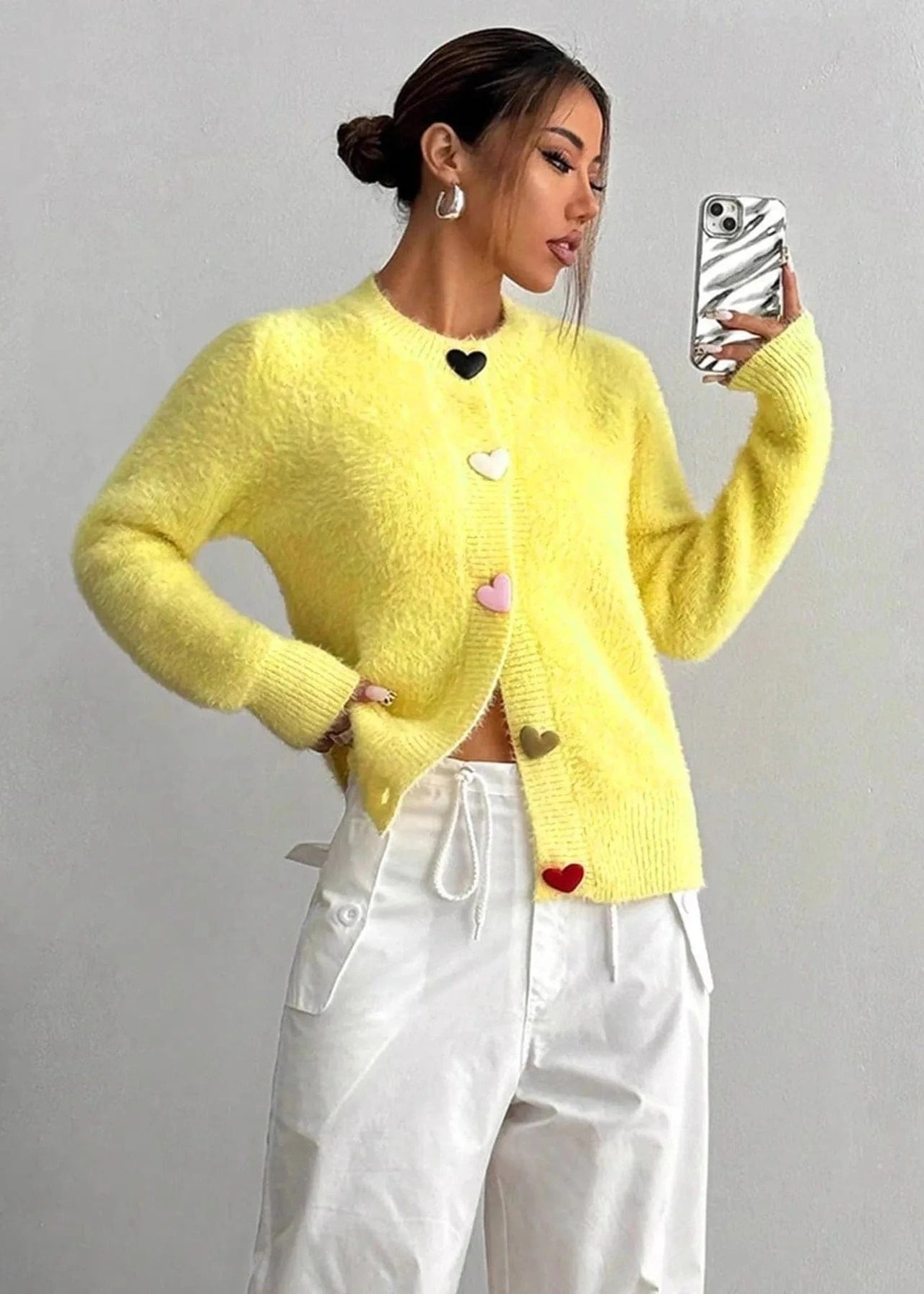 Woman wearing a yellow cardigan with heart patterns, taking a selfie against a plain background