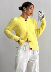 Woman wearing a yellow cardigan with heart patterns, taking a selfie against a plain background