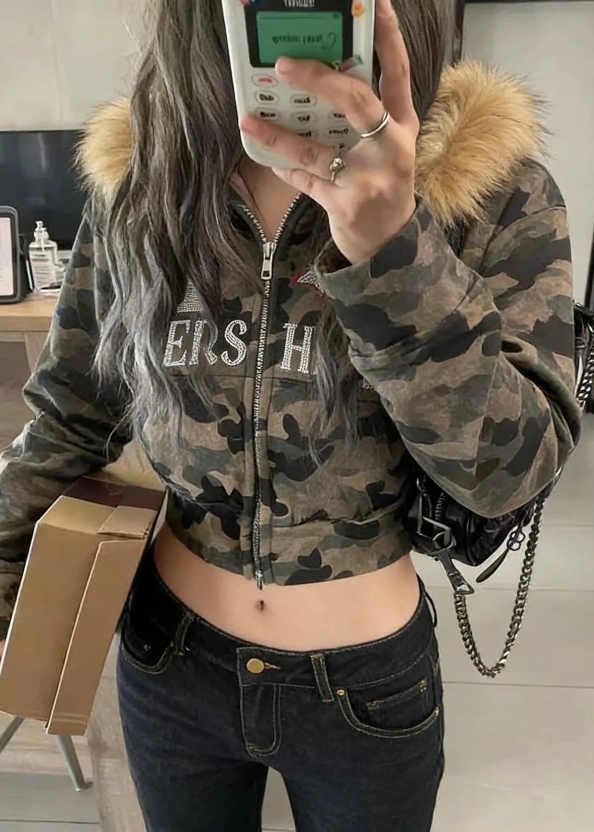Person wearing a camouflage jacket with fur hood, holding a phone and a box indoors.