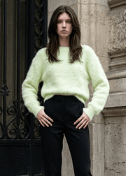 A model stands on stone steps wearing a fuzzy pale yellow-green long-sleeve sweater, black trousers, and black ankle boots.
