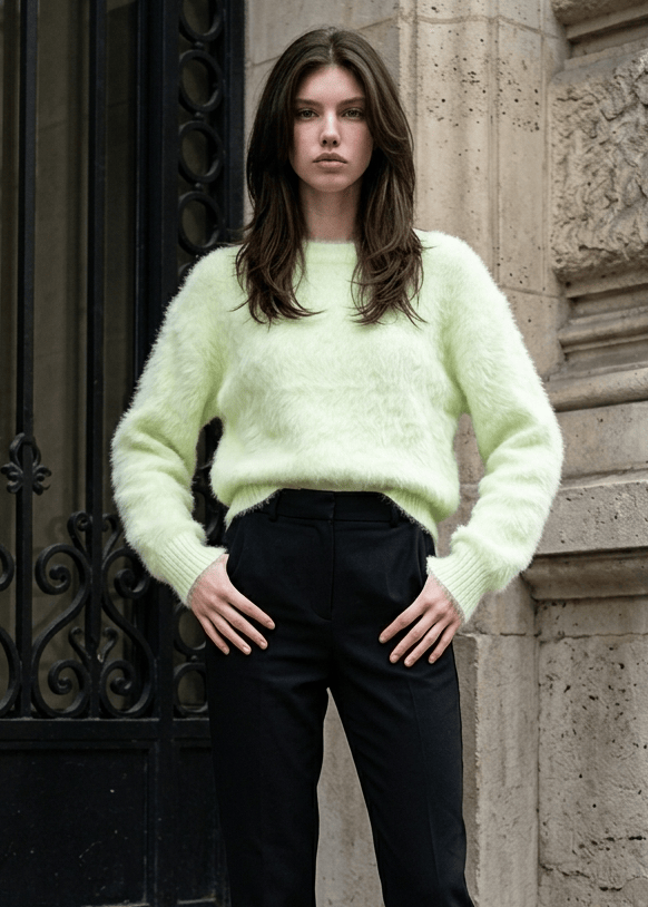 A model stands on stone steps wearing a fuzzy pale yellow-green long-sleeve sweater, black trousers, and black ankle boots.
