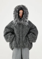 A model wearing a voluminous, oversized dark gray fuzzy faux-fur jacket with a large shaggy hood, paired with gray pants, shot against a plain light gray background.