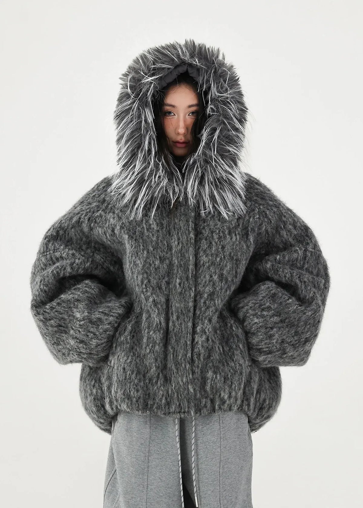 A model wearing a voluminous, oversized dark gray fuzzy faux-fur jacket with a large shaggy hood, paired with gray pants, shot against a plain light gray background.