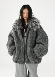 A model wearing a chunky, oversized gray faux fur jacket with a large shaggy gray and white faux fur collar, styled over a gray top and sweatpants against a plain white studio background.