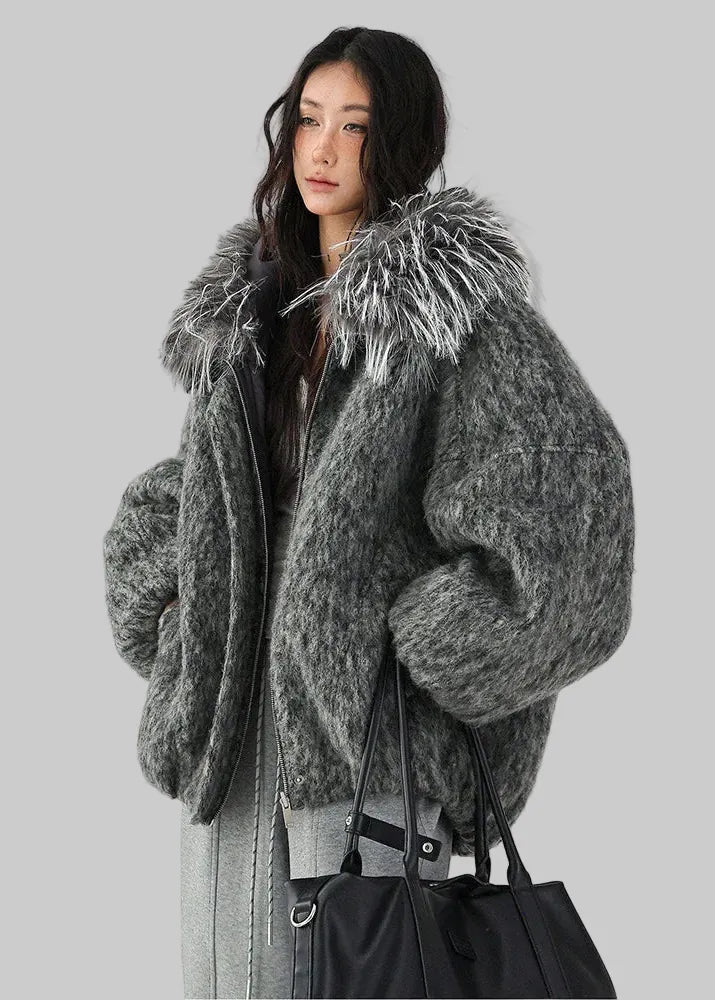 A woman wearing a voluminous, oversized fuzzy gray faux-fur jacket with a large shaggy faux-fur collar, styled with a black tote bag against a plain white studio background. 