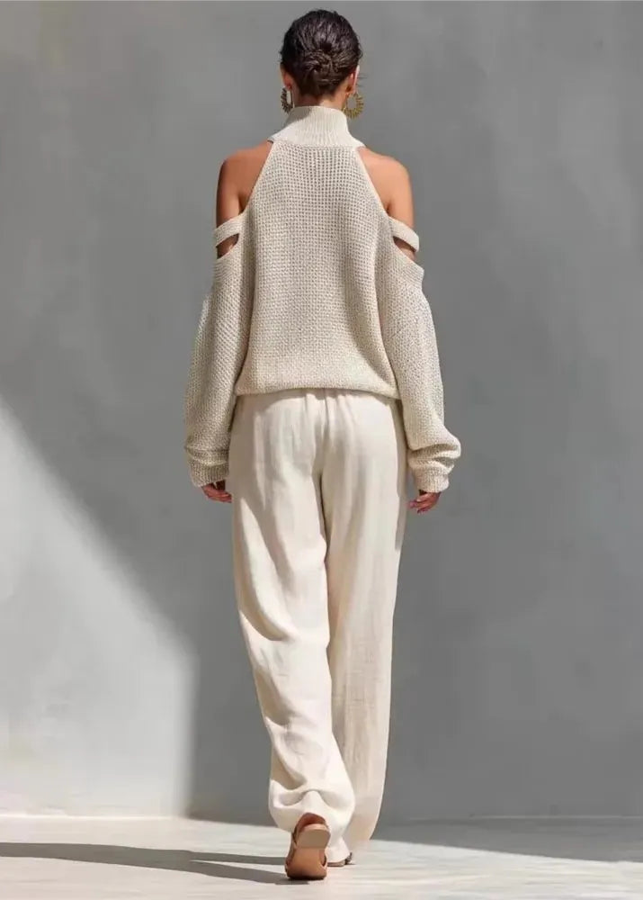 Person wearing a beige off-shoulder sweater and pants against a gray wall.