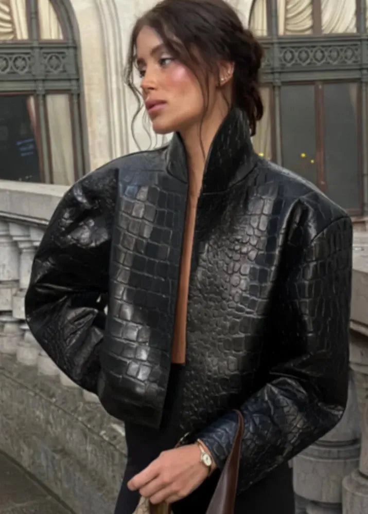 A woman wearing a black crocodile-embossed leather jacket with a high stand collar, photographed outdoors in front of a stone building.