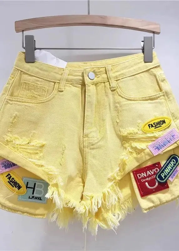 Yellow denim shorts with patches on a hanger against a gray background