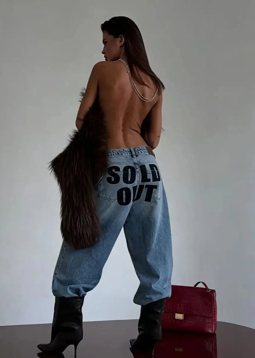A woman stands with her back to the camera wearing very loose light-wash blue jeans with 'SOLD OUT' printed in black across the back, black heeled boots, and holding a dark brown fur coat. A red handbag sits on the floor beside her against a plain light gray wall.