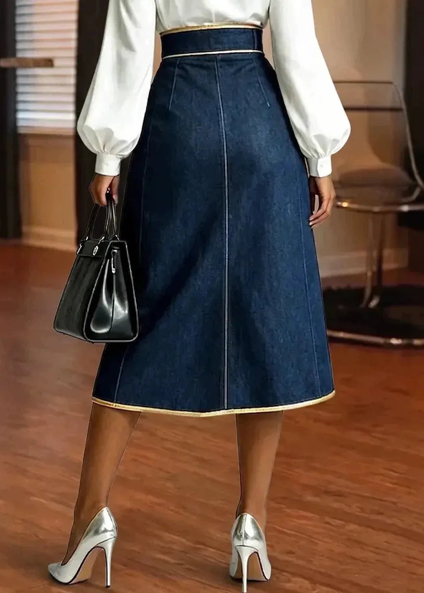 A woman wearing a dark blue denim midi skirt with gold trim, paired with a white long-sleeve blouse, silver high heels, and holding a black handbag, shown from the back in an indoor setting.