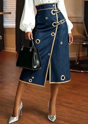A model wearing a dark blue denim midi skirt with gold trim, grommets, and belt details, paired with a white blouse, silver heels, and a black handbag in an indoor setting.