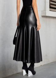 A woman wearing a high-waisted, black faux-leather flared midi skirt, paired with a black top, black socks, and chunky silver platform heels, standing against a gray concrete wall.
