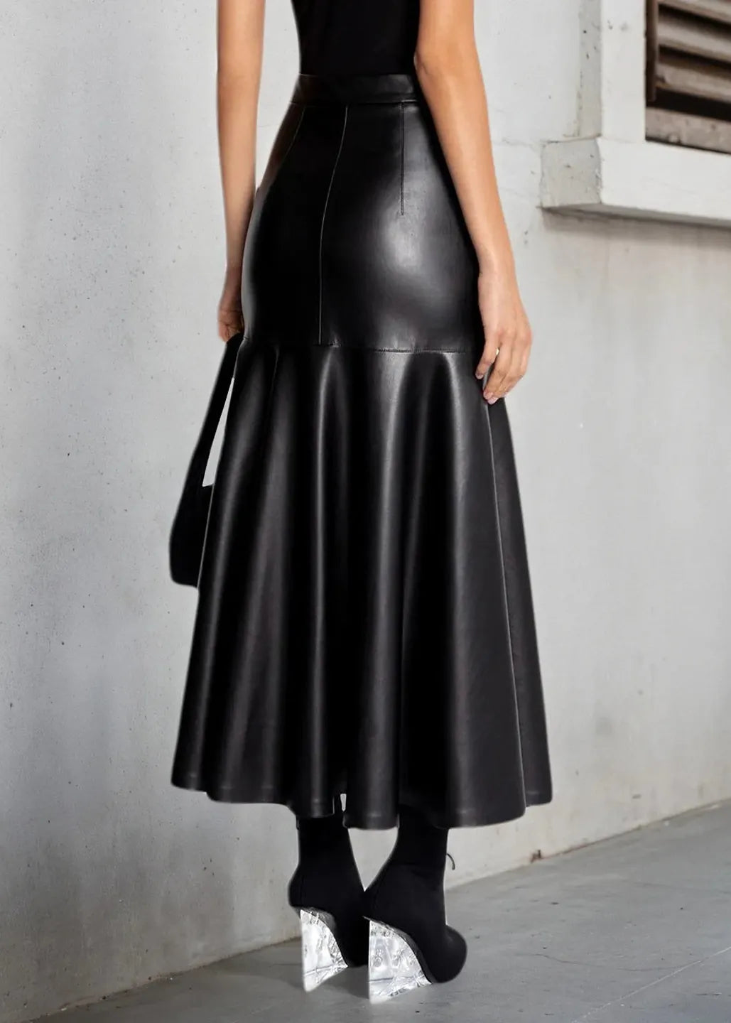 A woman wearing a high-waisted, black faux-leather flared midi skirt, paired with a black top, black socks, and chunky silver platform heels, standing against a gray concrete wall.