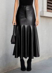 A model wearing a black faux-leather midi skirt with a tie waist, paired with a black bodysuit, black socks, and holding a black handbag against a gray concrete wall.