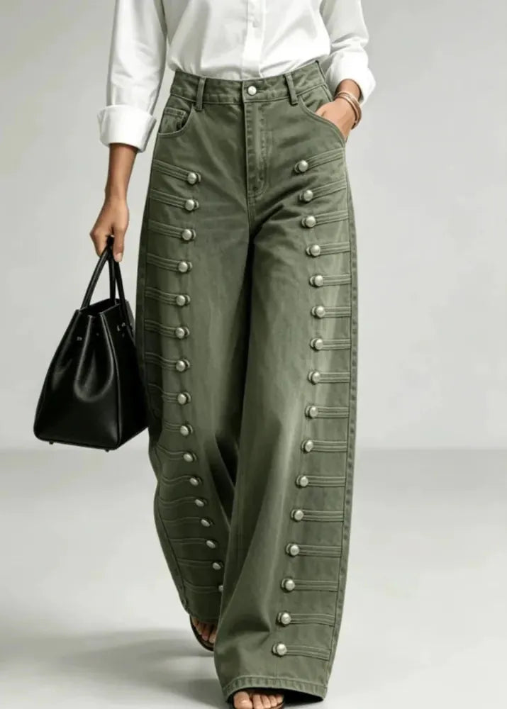 A model wearing high-waisted, wide-leg olive green pants with decorative silver button details down the sides, paired with a white shirt and holding a black handbag.