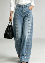 A person wearing light blue wide-leg denim jeans with decorative silver-tone button and strap detailing down the outer sides, styled with a white top and a black handbag against a light gray studio background.