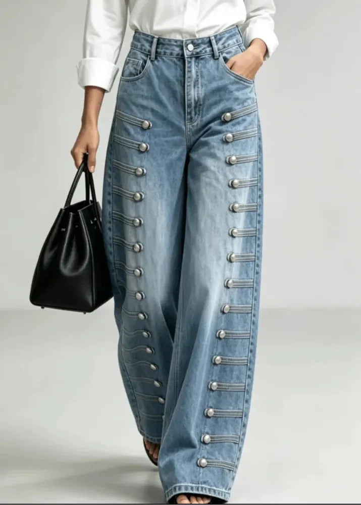 A person wearing light blue wide-leg denim jeans with decorative silver-tone button and strap detailing down the outer sides, styled with a white top and a black handbag against a light gray studio background.