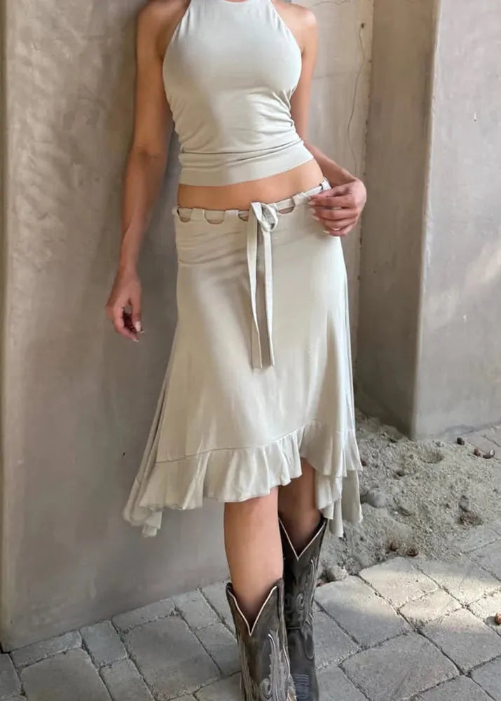 Person wearing a white top and skirt with cowboy boots standing against a concrete wall.