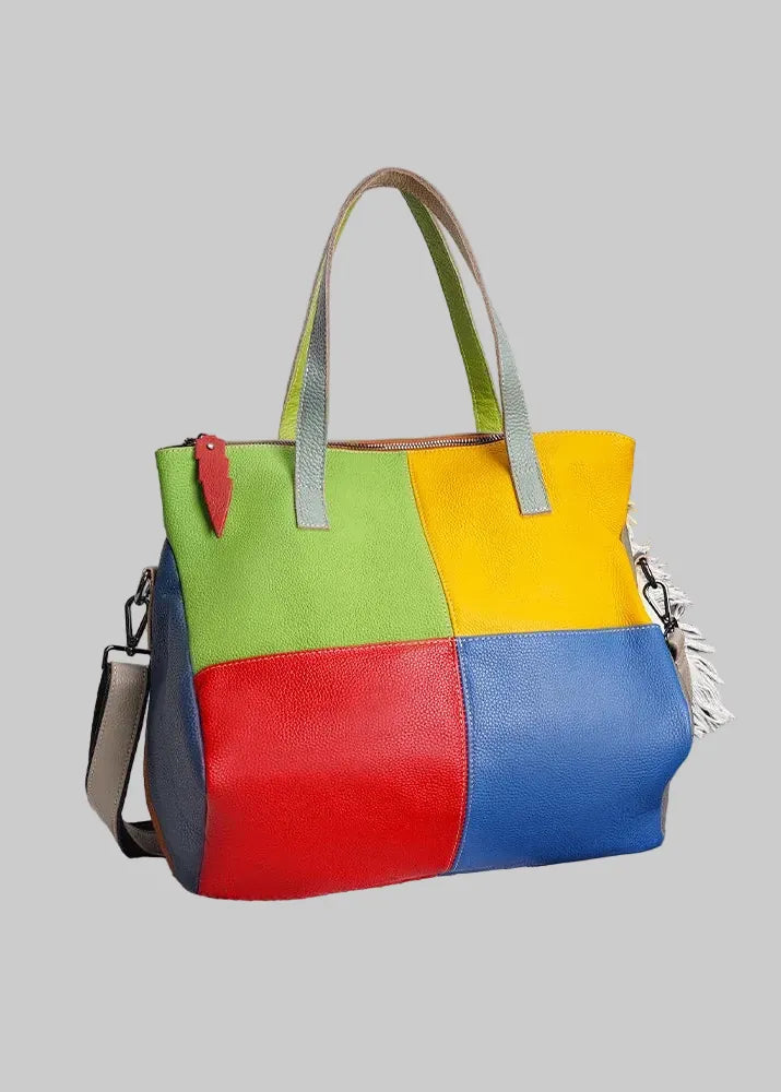 A Zency genuine leather handbag with a colorful patchwork design in green, yellow, red, and blue, featuring light gray handles, displayed on a dark leather sofa with a white throw blanket.
