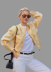 Woman in a yellow patterned jacket and white pants posing in front of a black gate.