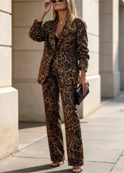 A woman wearing a leopard print blazer and matching wide-leg pants, standing outdoors on a sidewalk near beige stone columns. She is holding a small black clutch and wearing sunglasses.