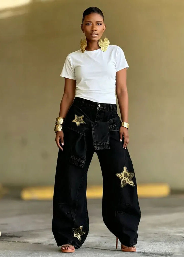 A woman stands in a concrete structure wearing oversized black denim pants with gold embroidered star patches, a white short-sleeve t-shirt, and gold statement earrings and bracelets.