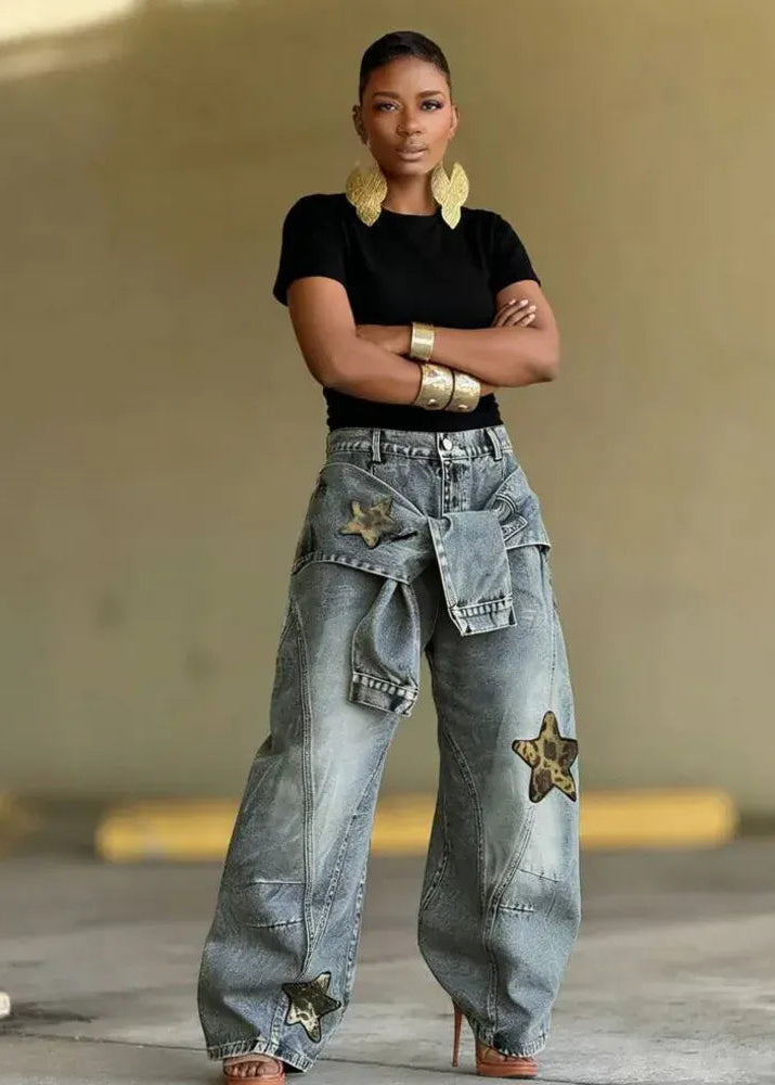 A model stands with arms crossed wearing light-wash wide-leg denim jeans featuring star patches and cargo pockets, a black short-sleeve top, and gold jewelry in an outdoor concrete setting.