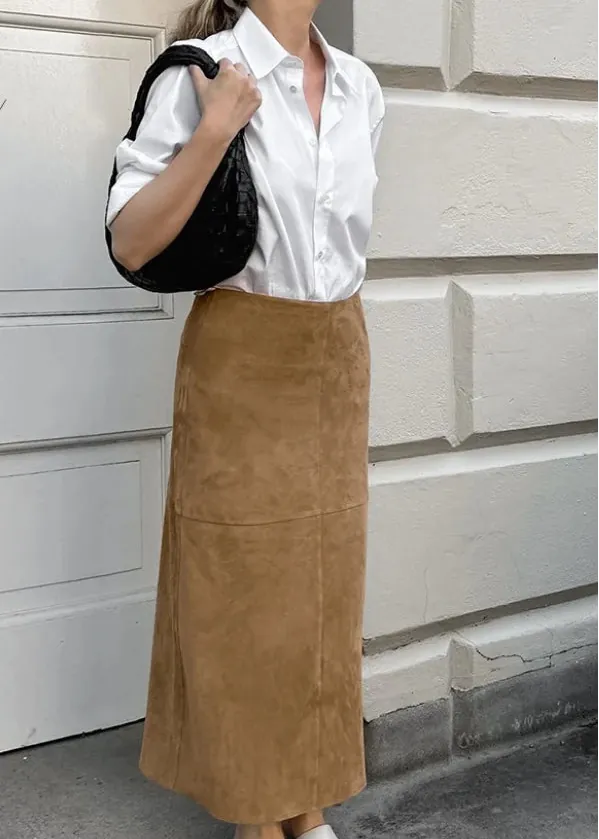 A woman wearing a white button-up shirt and a brown suede midi skirt, holding a black shoulder bag. She is standing outdoors against a white paneled door and textured wall.