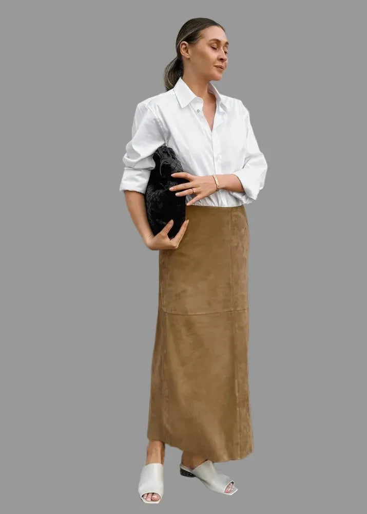 A woman stands outdoors against a light stone wall wearing a white button-up shirt, a long tan-brown suede-like skirt, white slide sandals, and holding a black clutch bag. The brand name 'SINGELIN' is visible in the top left corner.