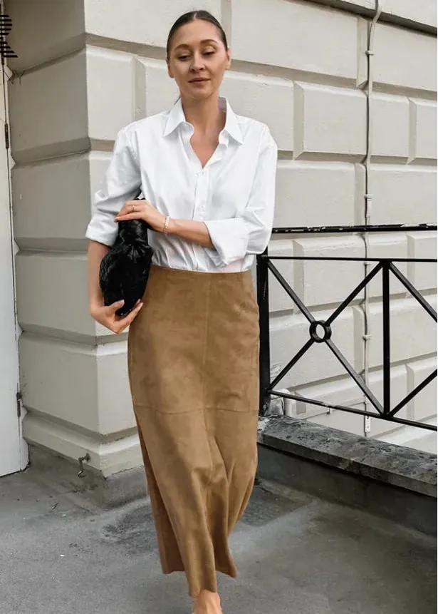A woman stands outdoors wearing a white button-up shirt tucked into a long tan skirt, with white slide sandals and a small black handbag. The background shows a light stone building wall, a white door, and a black metal railing.