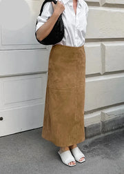 A woman wearing a long brown suede maxi skirt, a white button-down shirt, and white slide sandals, holding a black quilted shoulder bag. She is standing outdoors against a light-colored brick wall.