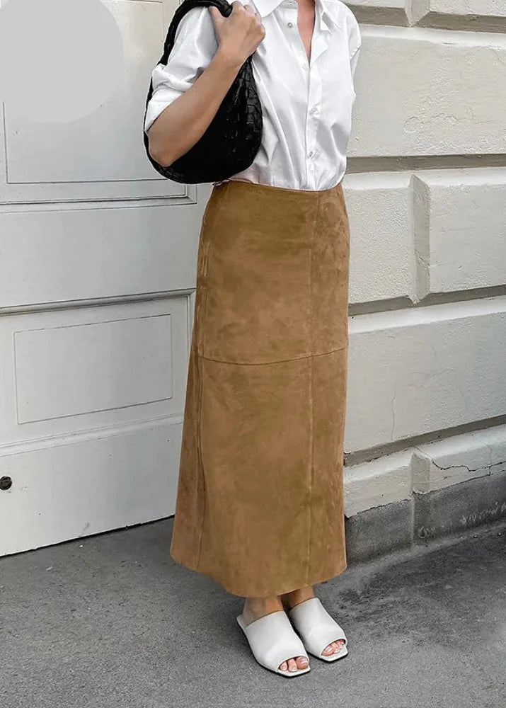 A woman wearing a long brown suede maxi skirt, a white button-down shirt, and white slide sandals, holding a black quilted shoulder bag. She is standing outdoors against a light-colored brick wall.