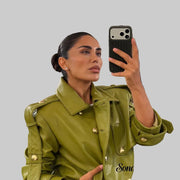 A woman takes a mirror selfie in a clothing boutique, wearing a structured olive green faux leather jacket with gold-tone buttons and shoulder epaulets. The boutique background shows racks of clothing.