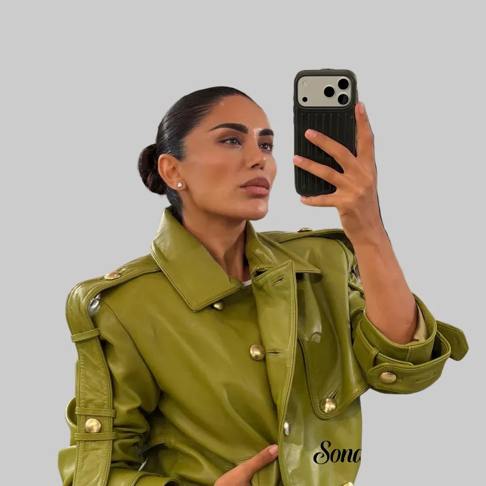 A woman takes a mirror selfie in a clothing boutique, wearing a structured olive green faux leather jacket with gold-tone buttons and shoulder epaulets. The boutique background shows racks of clothing.