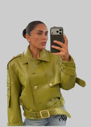 A woman takes a mirror selfie in a clothing store wearing a structured olive green faux-leather jacket with a matching belt and gold-tone buckle. The background shows clothing racks and store fixtures.