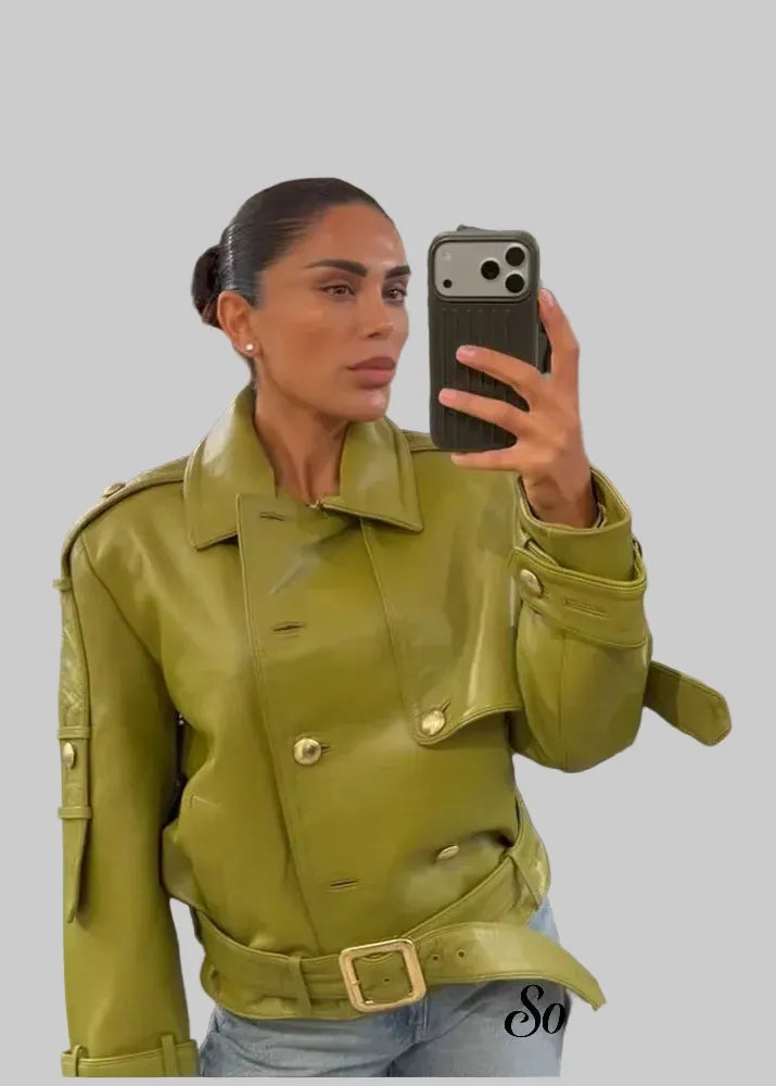A woman takes a mirror selfie in a clothing store wearing a structured olive green faux-leather jacket with a matching belt and gold-tone buckle. The background shows clothing racks and store fixtures.