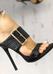 Black high-heeled sandal with a gold buckle worn by a person.