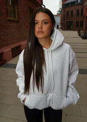 Woman wearing a gray hoodie standing on a tiled pavement with brick buildings in the background