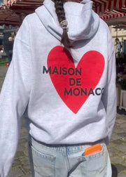 Person wearing a hoodie with a red heart and 'MAISON DE MONACO' text in an outdoor setting.