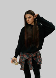 Woman standing against a brick wall, wearing a black sweater over a plaid shirt.