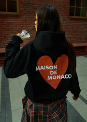 Person wearing a black hoodie with a red heart and 'MAISON DE MONACO' text, standing in front of a brick wall.
