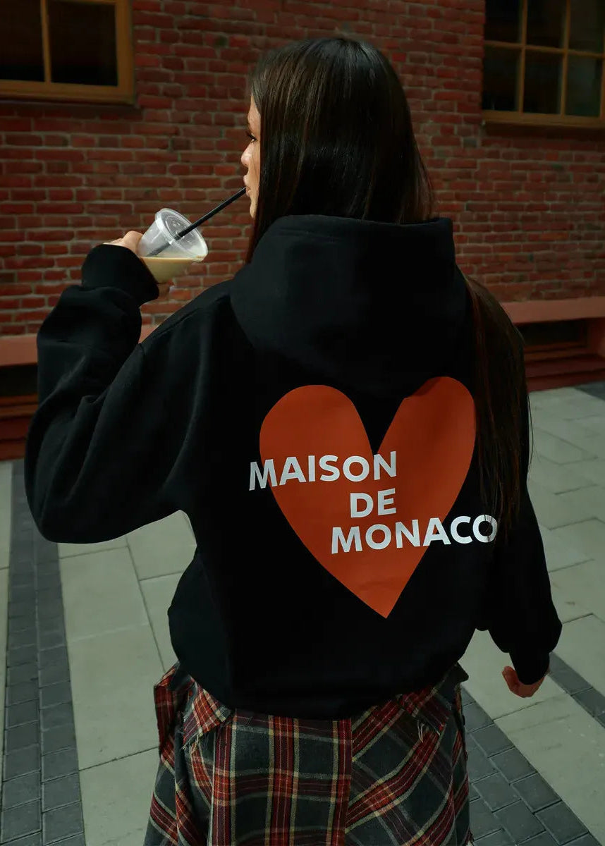Person wearing a black hoodie with a red heart and 'MAISON DE MONACO' text, standing in front of a brick wall.