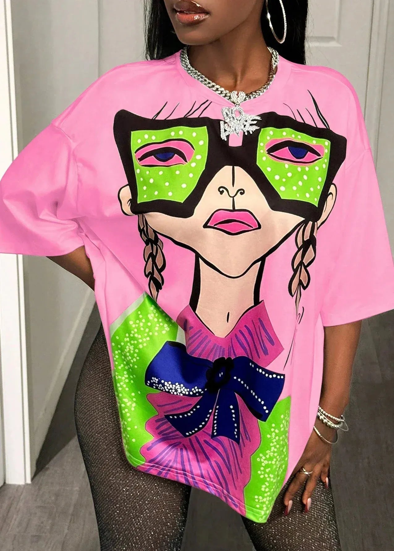 Person wearing a pink t-shirt with a colorful graphic design of a face with green sunglasses and a bow.