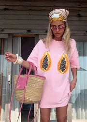 Person wearing a pink dress with papaya graphic, holding a straw bag, standing in front of a wooden door.