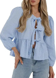 Woman wearing a blue checkered blouse with long sleeves and tied details, paired with light blue jeans on a white background.