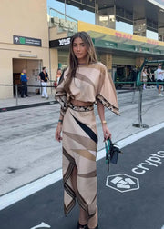 A woman stands outdoors on a street or event area wearing a beige and brown geometric print two-piece outfit with a cropped top and matching skirt. She is holding a small black handbag, with buildings, signage, and other people visible in the background.