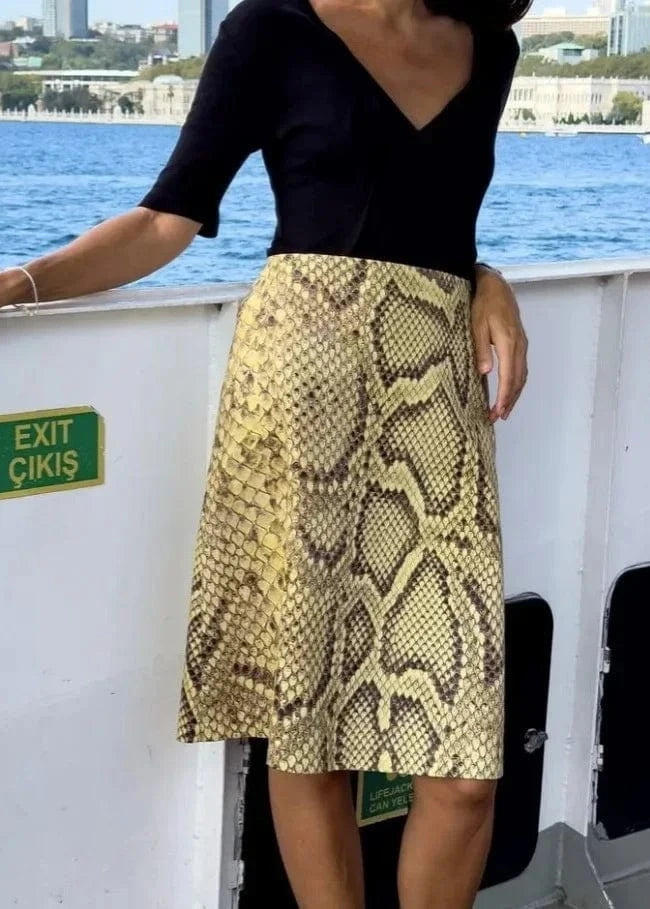 Person wearing a black top and snake print skirt on a boat with water and cityscape in the background.